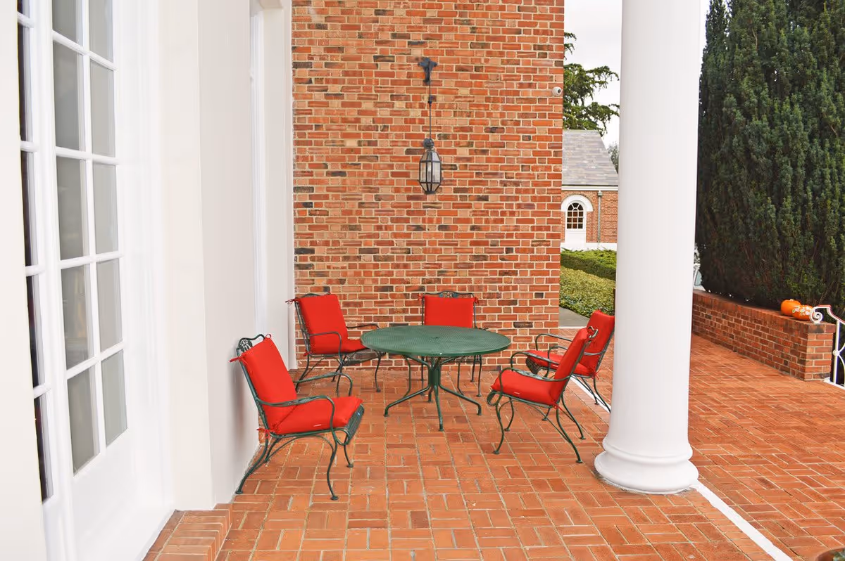 Outdoor patio area with a round green metal table surrounded by four green metal chairs with red cushions. The patio has a brick floor and a brick wall with a lantern-style light fixture. There is a large white column on the right side and some greenery in the background.