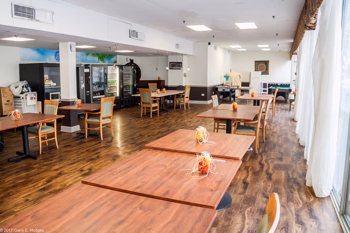 Spacious dining/activity room with wooden tables and chairs, vending machines along the back wall, and large windows with curtains on the right.