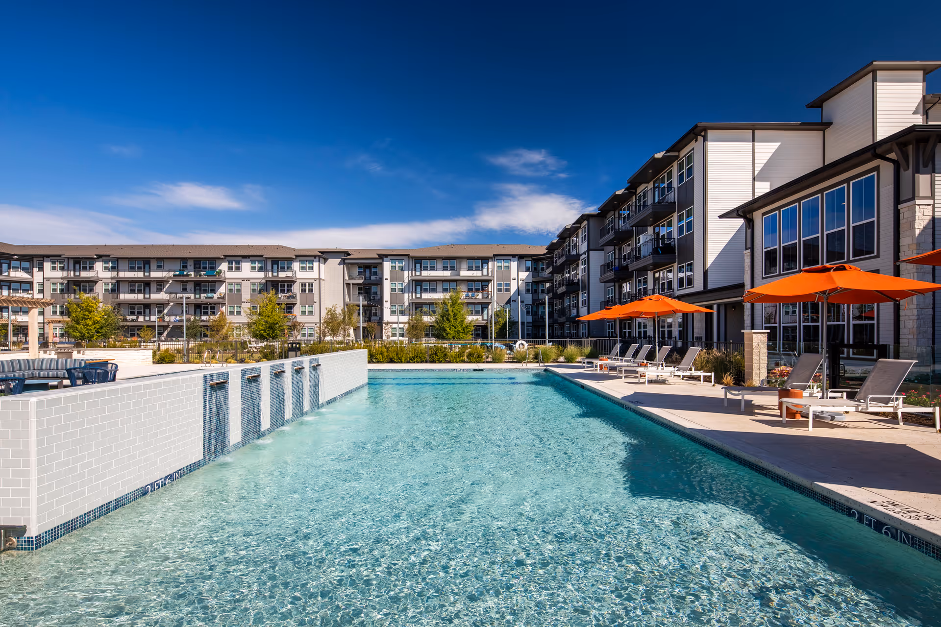 Outdoor swimming pool with lounge chairs and orange umbrellas beside a multi-story residential building under a blue sky.