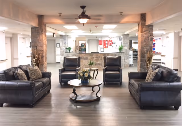 A spacious and well-lit common area with two dark leather sofas and two matching armchairs arranged around a round wooden coffee table with a flower arrangement. The room features stone pillars, wood flooring, and a ceiling fan with a light fixture. In the background, there is a reception desk with plants and various notices on the walls.