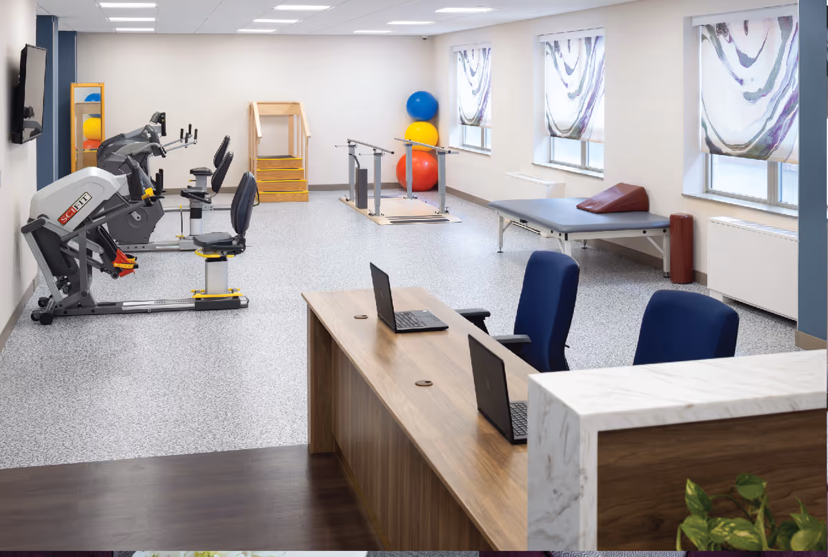 A bright rehabilitation or physical therapy room with exercise equipment including stationary bikes, therapy stairs, parallel bars, and large colorful exercise balls. There is a wooden desk with two laptops and blue chairs in the foreground, and windows with patterned curtains letting in natural light.
