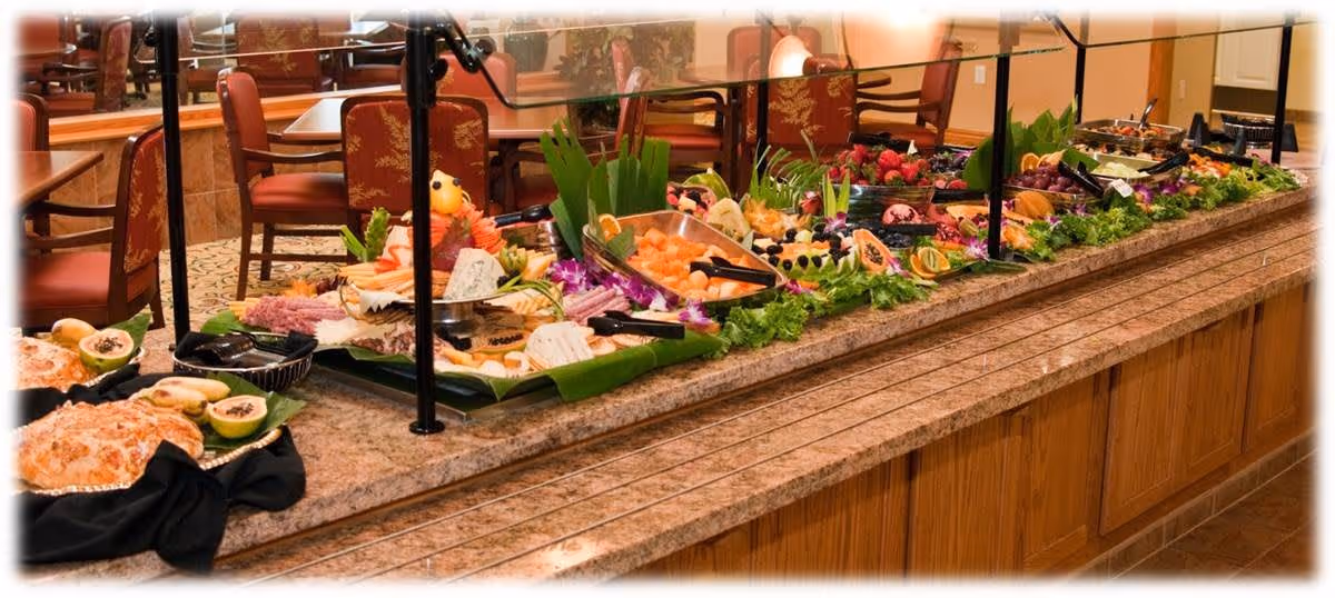 A buffet-style food serving area with a variety of fresh fruits, vegetables, cheeses, and other dishes arranged on a granite countertop. There are serving utensils and plates available, with chairs and tables visible in the background.