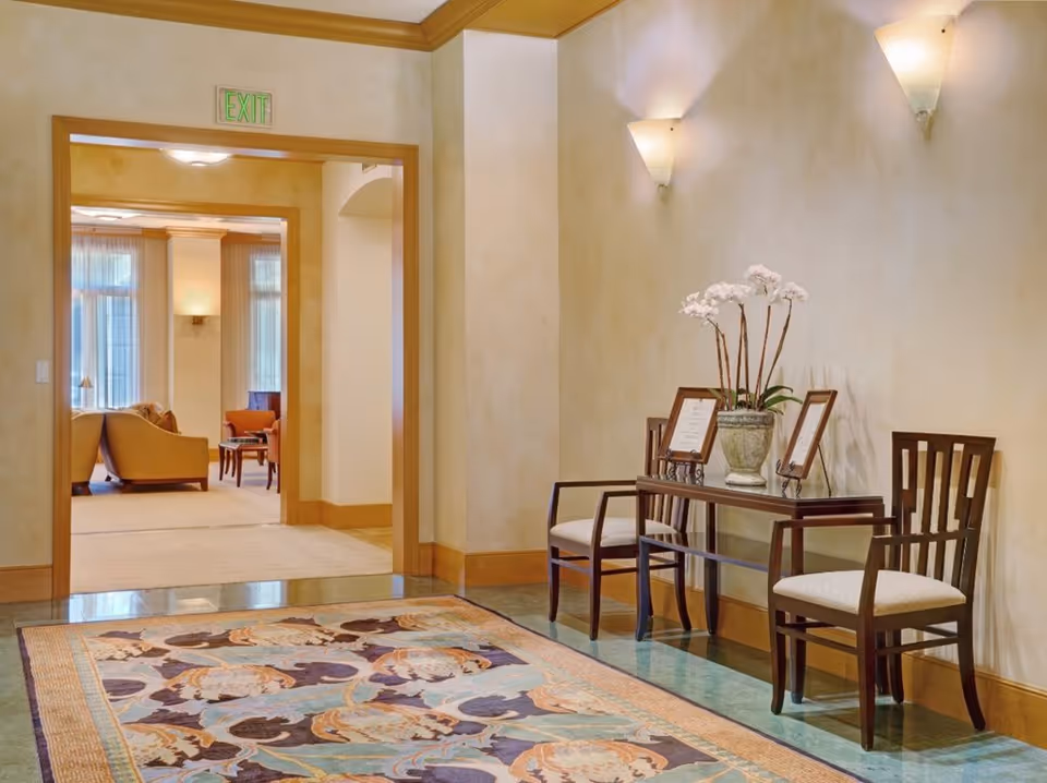 A well-lit interior hallway with beige walls and wooden trim. Two wooden chairs with light cushions flank a glass-top table holding a potted orchid and two framed items. A colorful patterned rug covers part of the floor. Through an open doorway, a living area with armchairs and large windows with sheer curtains is visible. An illuminated exit sign is above the doorway.