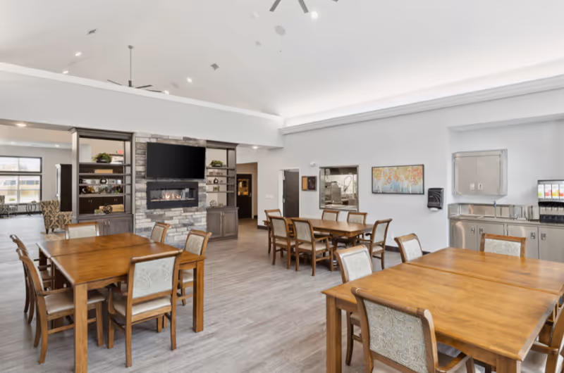 A spacious senior living dining area with several wooden tables and chairs arranged neatly. The room features a stone fireplace with a mounted flat-screen TV above it, built-in shelves with decorative plants, and a beverage station with a sink and drink dispensers. The space has light-colored walls, a high ceiling with ceiling fans, and large windows allowing natural light.