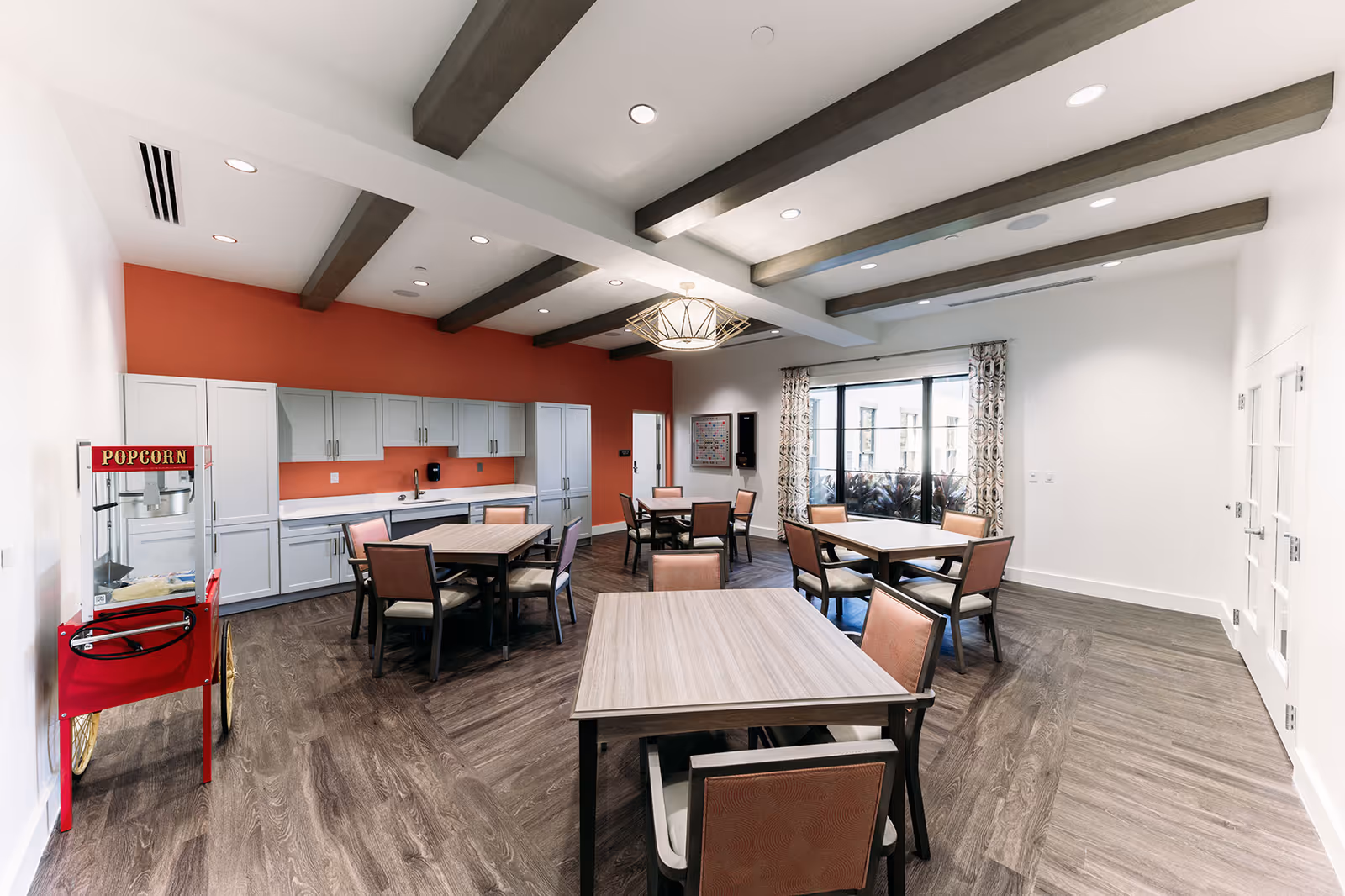 Bright communal dining/activity room with multiple tables and chairs, a kitchenette along an orange accent wall, and a popcorn machine.