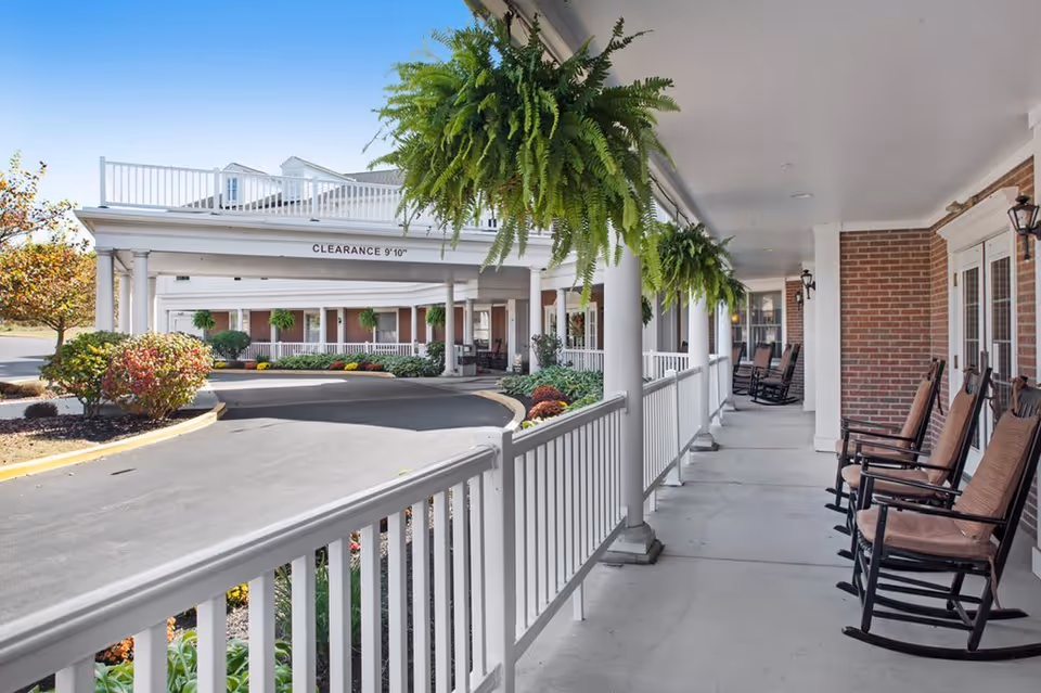 Covered porch area of Poland Village Assisted Living with several cushioned rocking chairs lined up along a white railing. Hanging green ferns are suspended from the ceiling. The driveway curves around landscaped bushes and flowers, with a clearance sign of 9 feet 10 inches visible above the entrance.