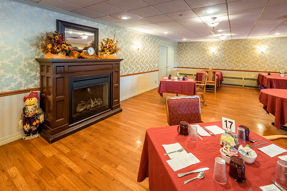 A dining room with several tables covered in red tablecloths, each set with utensils, cups, and menus. The room has wooden flooring, wallpapered walls with a floral pattern, and a dark wooden fireplace decorated with autumn-themed floral arrangements and a clock. A small scarecrow decoration stands next to the fireplace.
