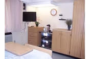 A small, tidy room with light wood furniture including a dresser, a cabinet, and a bed with a wooden headboard. A flat-screen TV is mounted on the wall above the dresser. There is a clock on the wall, a small plant on the cabinet, and a coffee or tea station with a pot and cups on a small shelf unit. The room has light-colored curtains and a window air conditioning unit.