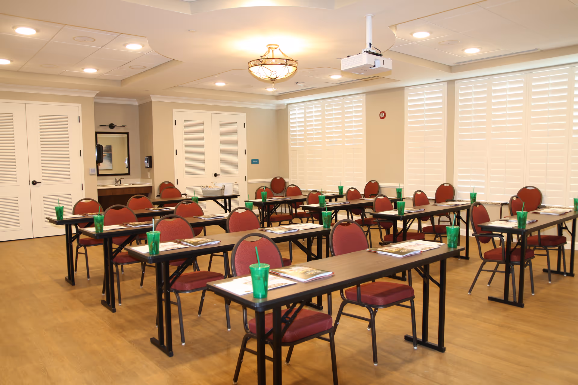 Bright multipurpose activity room set up with rows of tables, red chairs, green cups, and a ceiling projector.