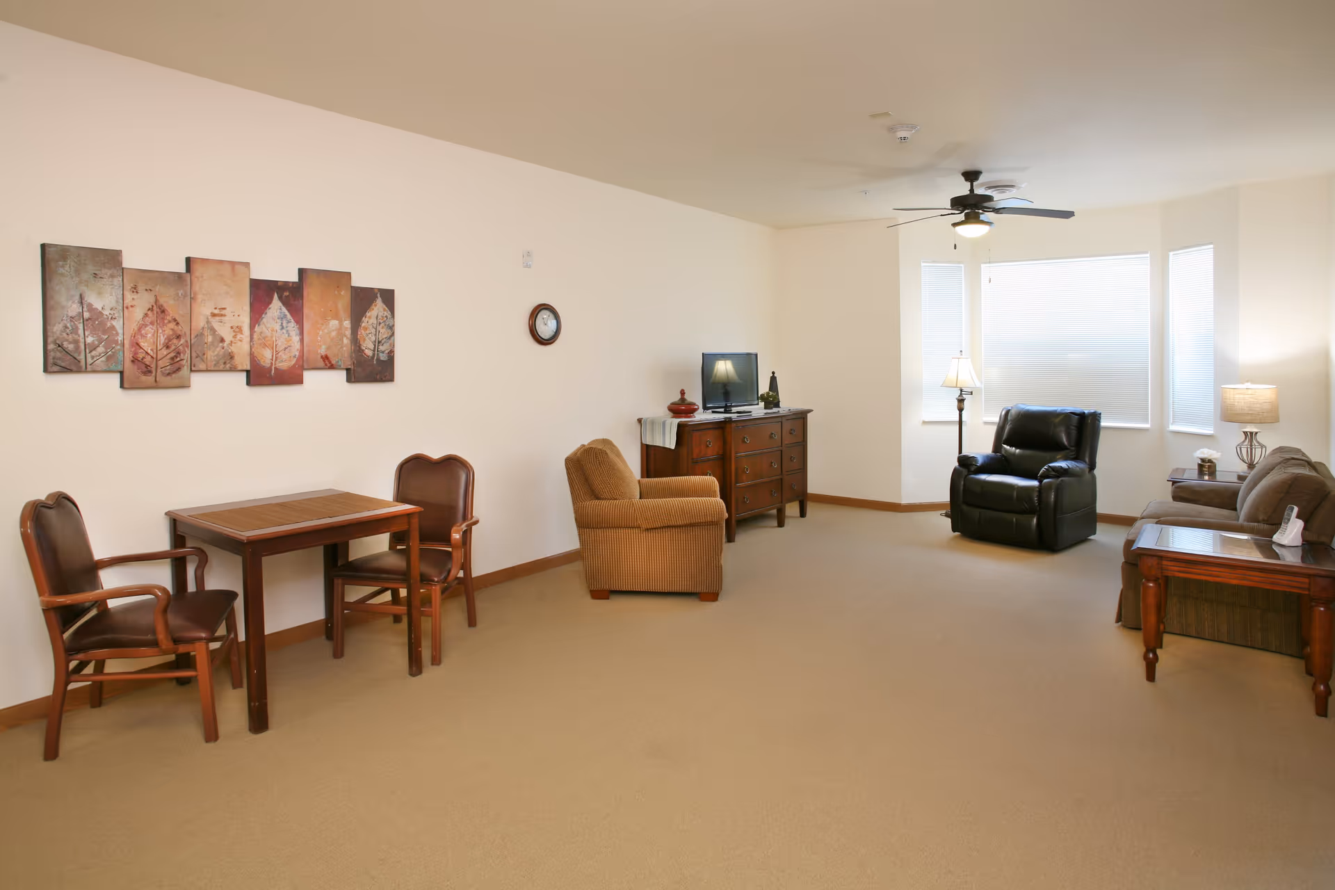 A spacious living room with beige carpet and cream-colored walls. The room features a small wooden table with two chairs on the left, a brown armchair, a wooden dresser with a small TV and decorative items, a black recliner near a bay window with blinds, a brown sofa, and a wooden side table with a lamp and tissue box. A ceiling fan with a light is mounted on the ceiling. There is a multi-panel wall art with leaf designs and a round clock on the wall.
