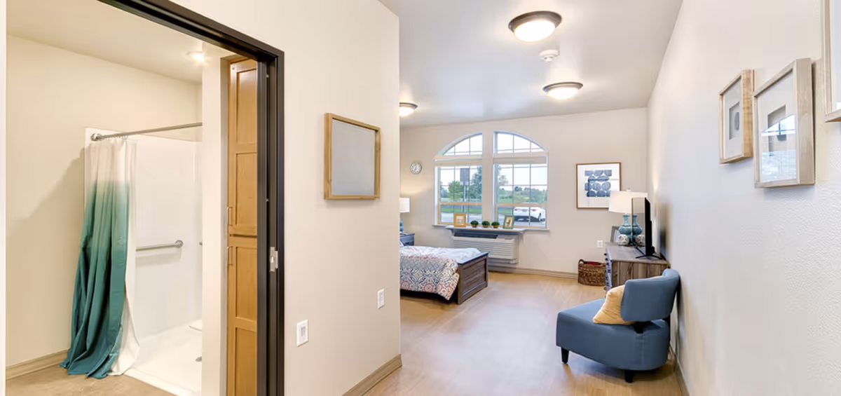 Interior view of a senior living facility room at Windsong at Northridge showing a bedroom area with a bed, a blue armchair with a pillow, a dresser with a TV and lamps, and a window with a scenic outdoor view. To the left, there is a bathroom entrance with a shower area visible, featuring a white and teal shower curtain.