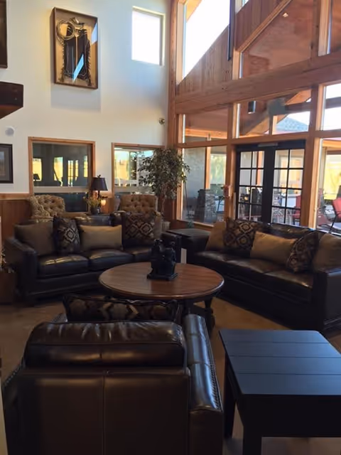 A cozy living room area with two dark leather sofas, a matching leather armchair, and a round wooden coffee table in the center. The room features large windows with wooden frames letting in natural light, a tall plant in the corner, and a high ceiling with wooden beams. There are decorative pillows on the sofas and framed artwork on the walls.