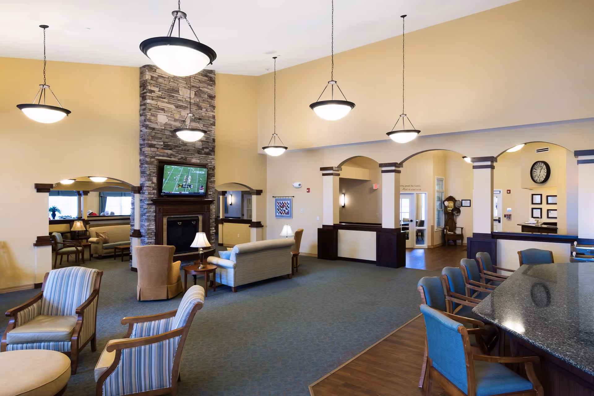 A spacious and well-lit living room area in a senior living facility with beige walls and blue carpeting. The room features multiple seating arrangements including armchairs and sofas around a stone fireplace with a mounted TV above it. There are several hanging pendant lights and a large clock on the wall. The room has arched doorways and dark wood trim accents, with a granite countertop bar area with blue cushioned chairs on the right side.