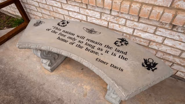 A curved concrete bench with military insignias and a quote engraved on the seat. The quote reads, 'This nation will remain the land of the free only so long as it is the home of the brave.' - Elmer Davis. The bench is placed on a concrete surface next to a brick wall.