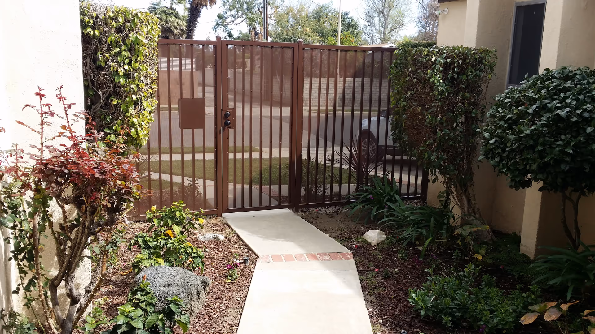 A small outdoor garden area with a concrete pathway leading to a brown metal gate. The garden has various green plants and shrubs on both sides of the pathway. The gate opens to a sidewalk and a parked car is visible in the background.