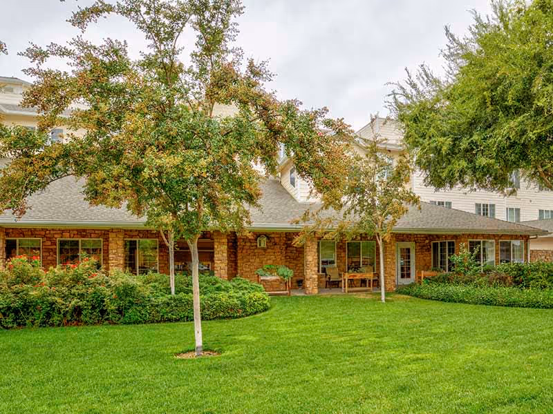 Covered stone-front porch of a senior living building with a green lawn, trees, and outdoor seating.