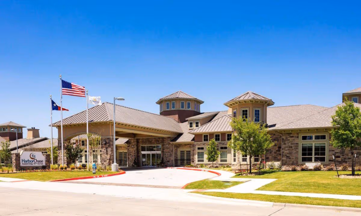 Exterior view of a senior living facility building with a stone facade, multiple peaked roofs, and two small towers. There are three flagpoles with the American flag, Texas state flag, and a facility flag. The entrance has a covered drop-off area and a sign that reads HarborChase Assisted Living - Memory Care. The surrounding area includes a well-maintained lawn, trees, and a clear blue sky.