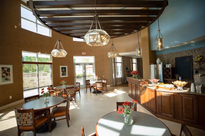 A bright dining area in a senior living facility with round tables and chairs, each table decorated with a small vase of red flowers. Large windows allow natural light to fill the room. The ceiling has exposed wooden beams and modern pendant lights. A wooden counter with a granite top holds baskets of bread and a beverage dispenser. The walls are painted in warm tones with some artwork and a chalkboard on one side.