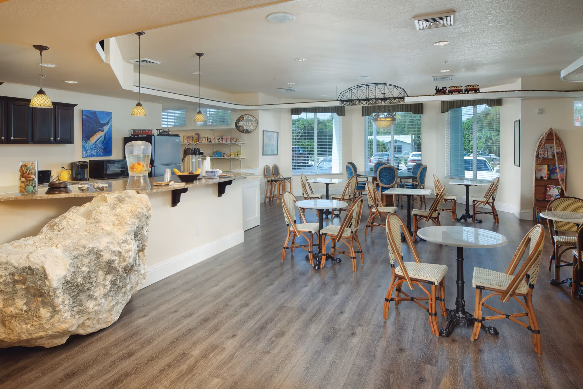 Bright communal dining area with multiple small round tables and chairs, a service counter with pendant lights and a large decorative rock at the counter.