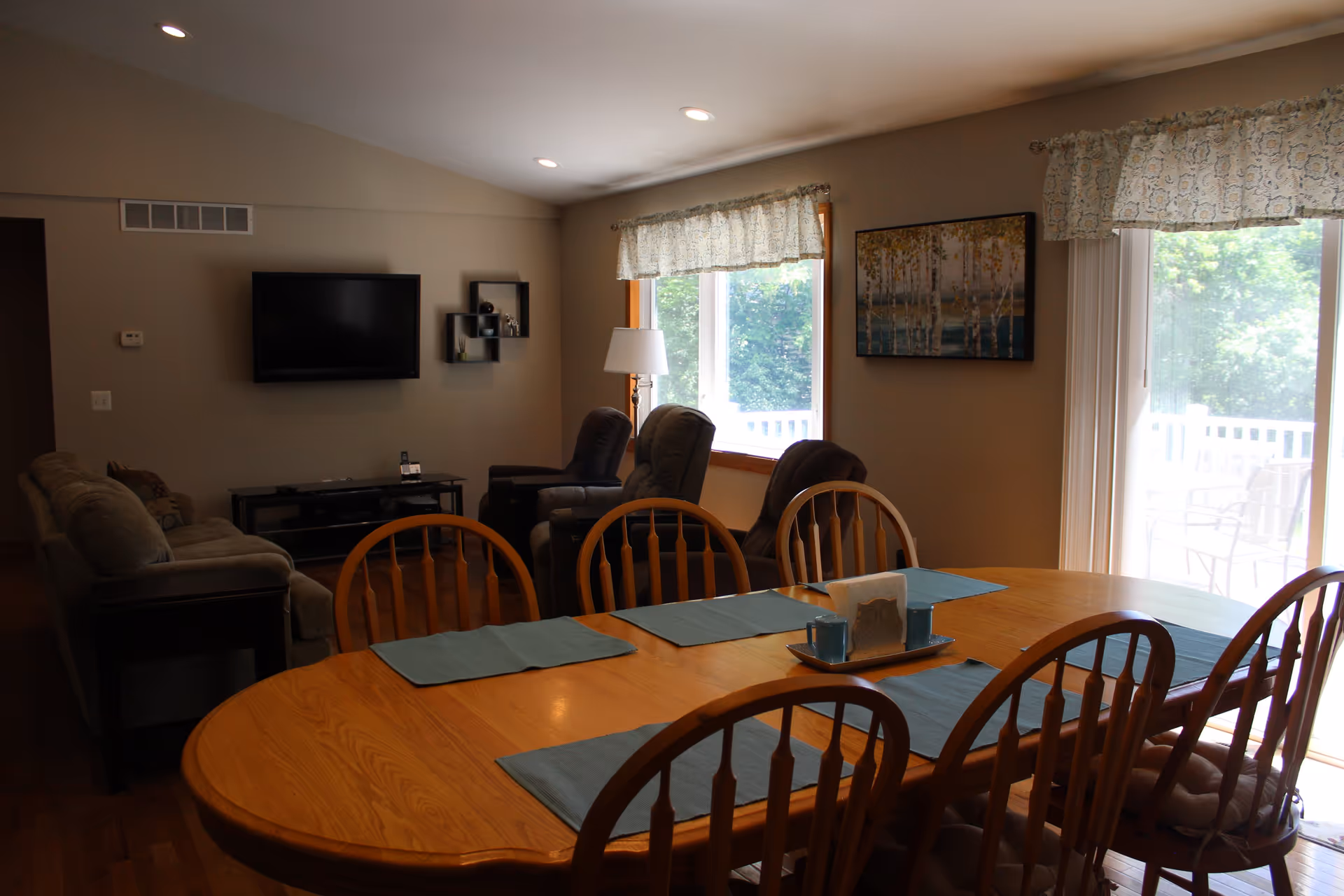 A cozy living and dining area with a wooden dining table set with blue placemats and a centerpiece. The living room has a sofa, three recliner chairs, a wall-mounted flat-screen TV, and a window with patterned curtains letting in natural light. A sliding glass door leads to an outdoor patio with chairs.