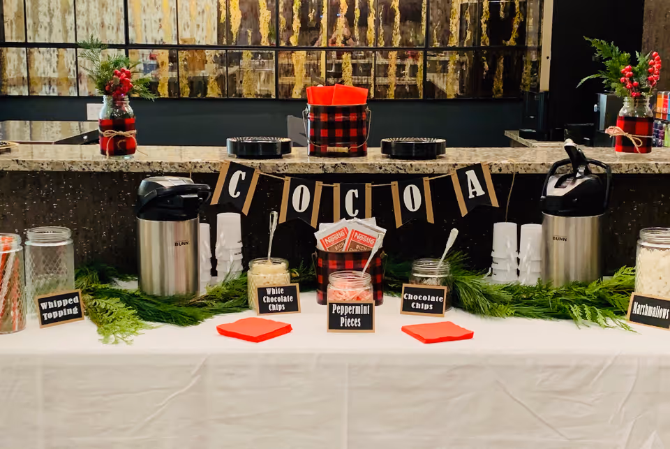 Festive hot cocoa bar with beverage dispensers, labeled toppings, cups, and holiday decorations on a countertop.