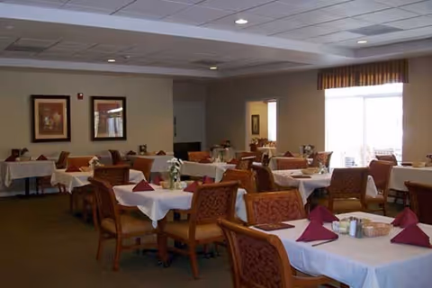 A dining room with multiple tables covered in white tablecloths and set with folded burgundy napkins, salt and pepper shakers, and small flower arrangements. The room has beige walls, framed artwork, and a large window with striped curtains letting in natural light.