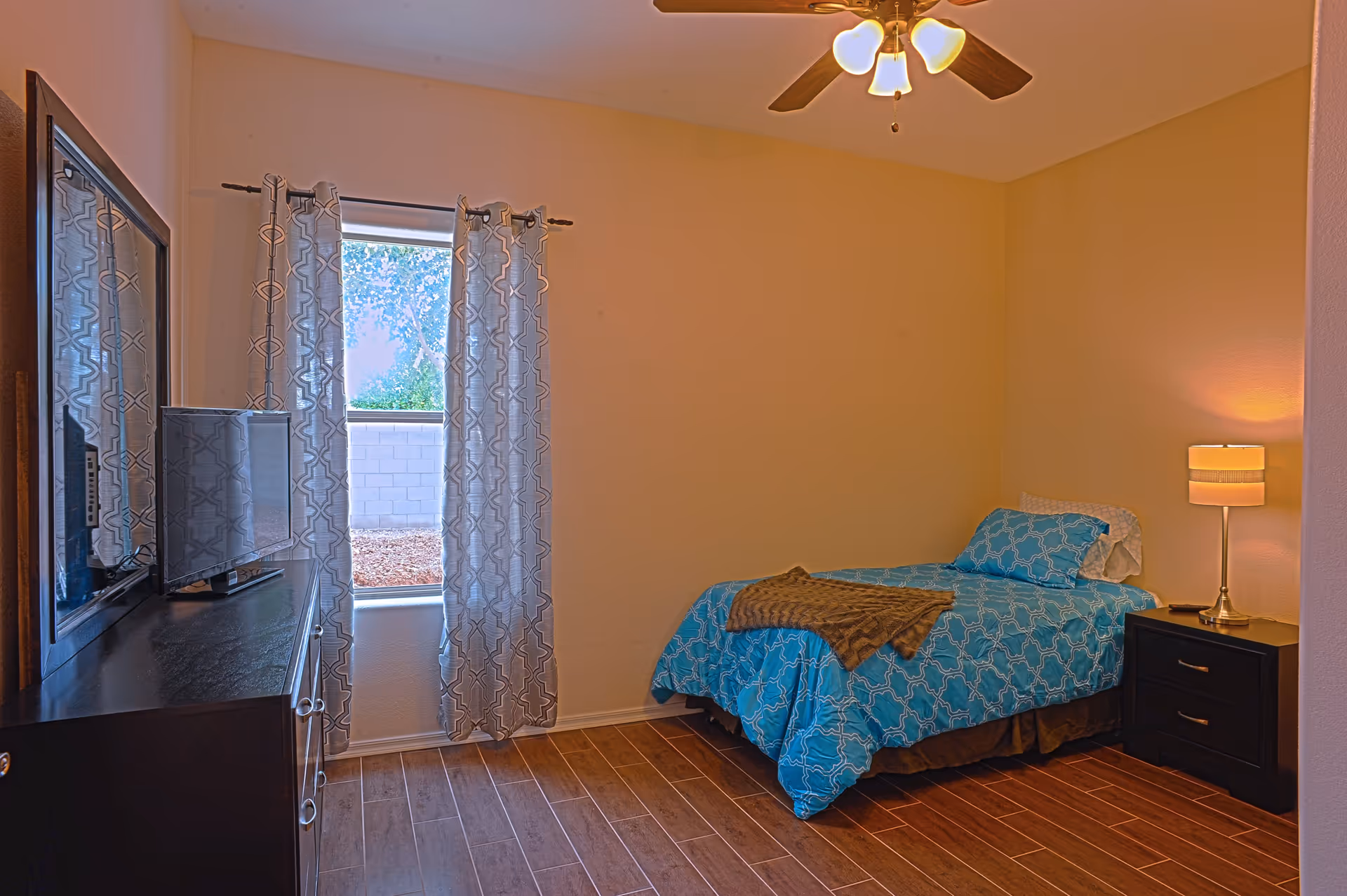A small bedroom with a single bed covered in blue patterned bedding and a brown throw blanket. Next to the bed is a black nightstand with a lamp and a remote control. Across from the bed is a black dresser with a flat-screen TV on top. A window with patterned curtains lets in natural light, and a ceiling fan with lights is mounted on the ceiling. The floor is wooden with a warm tone.