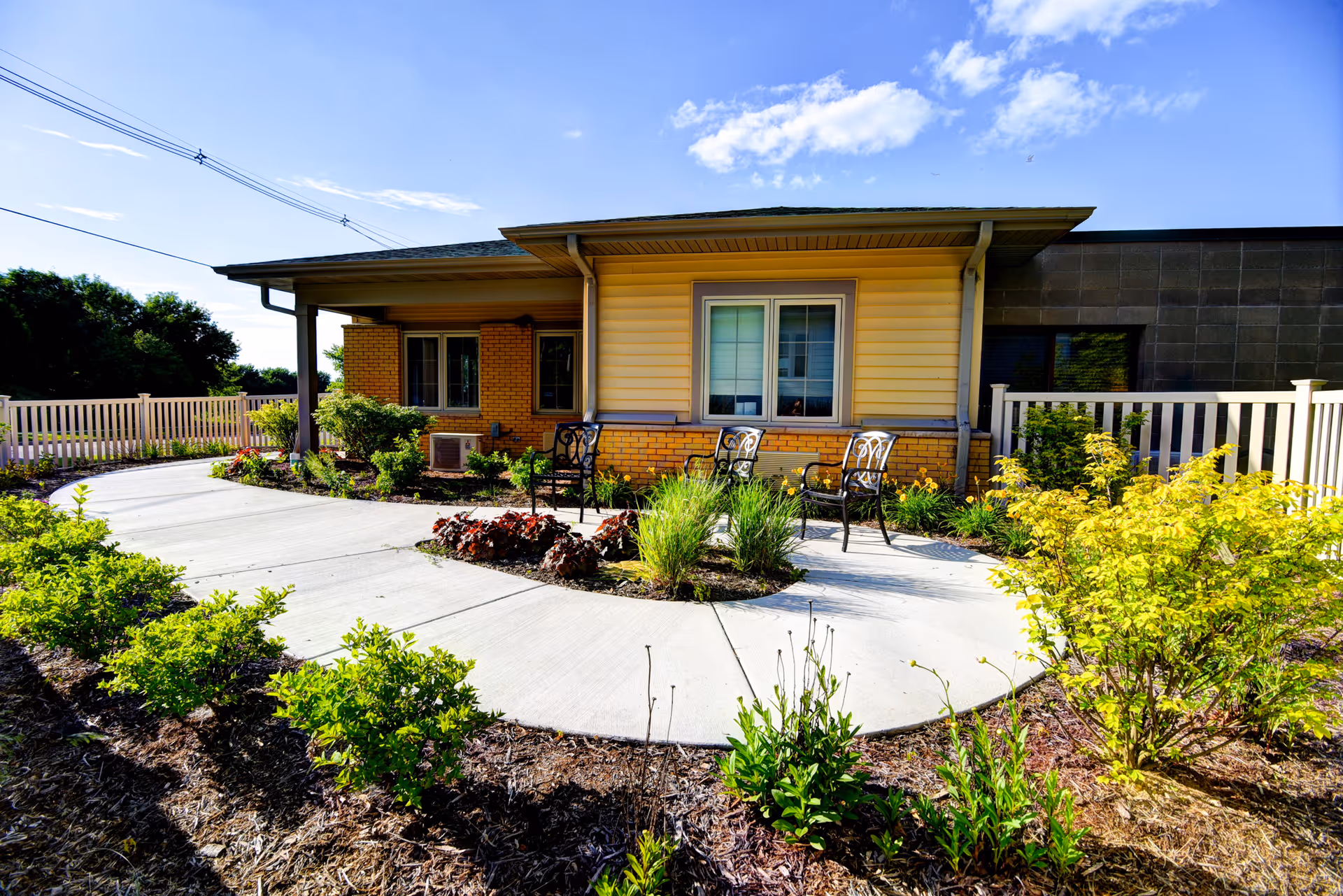 Sunlit single-story building exterior with a curved concrete walkway, landscaped beds and metal patio chairs in front of a window.