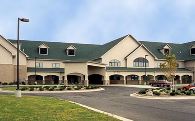 Exterior view of a large two-story retirement center building with a green roof, multiple windows, and an arched entrance. The building is surrounded by a driveway, landscaped areas with grass, small trees, and shrubs, and a few parked cars.