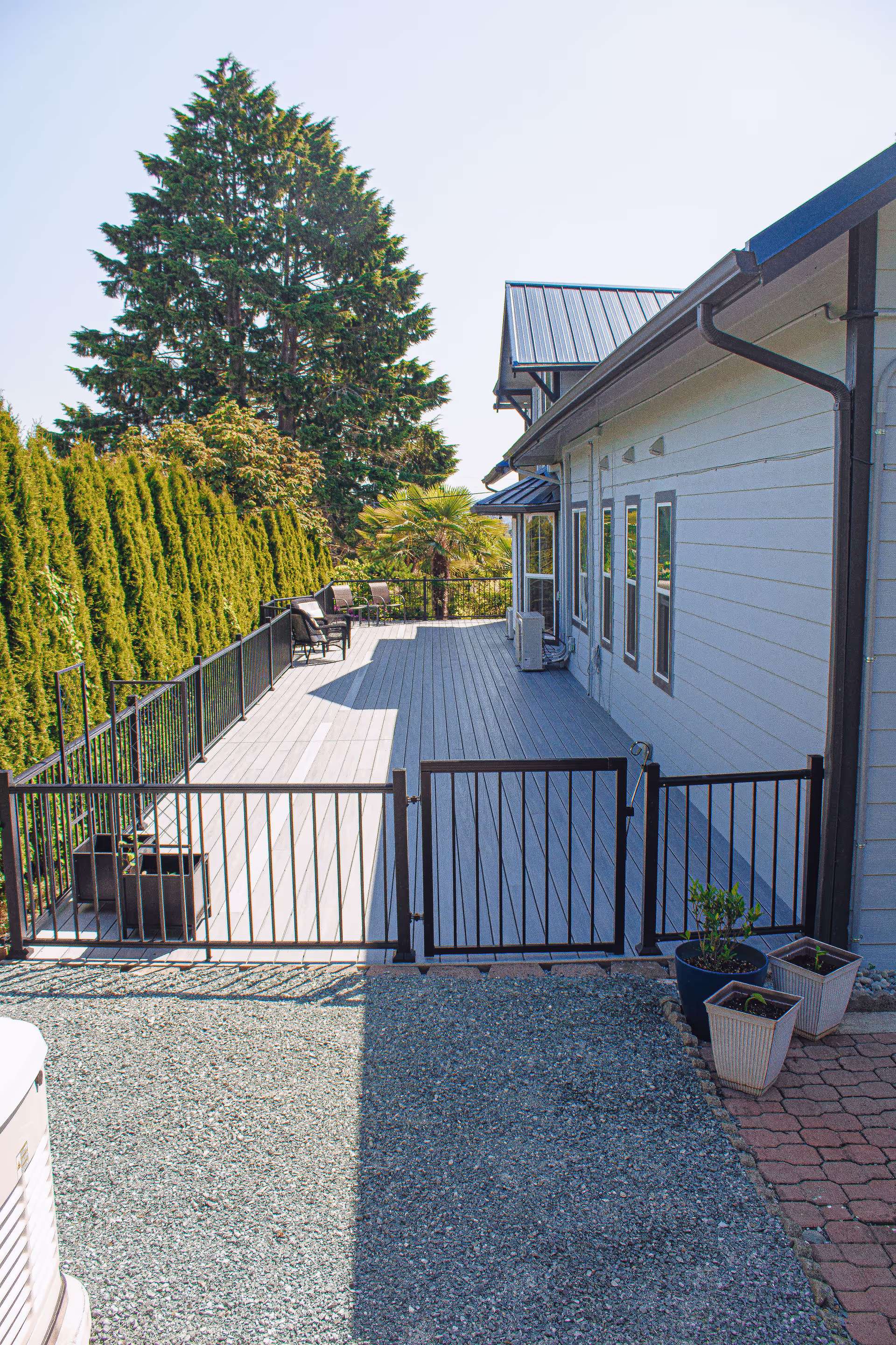 A fenced wooden deck alongside a light-colored house with chairs and tall trees in the background.
