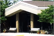Covered entrance to a building with glass double doors, brick columns, benches, and potted plants.