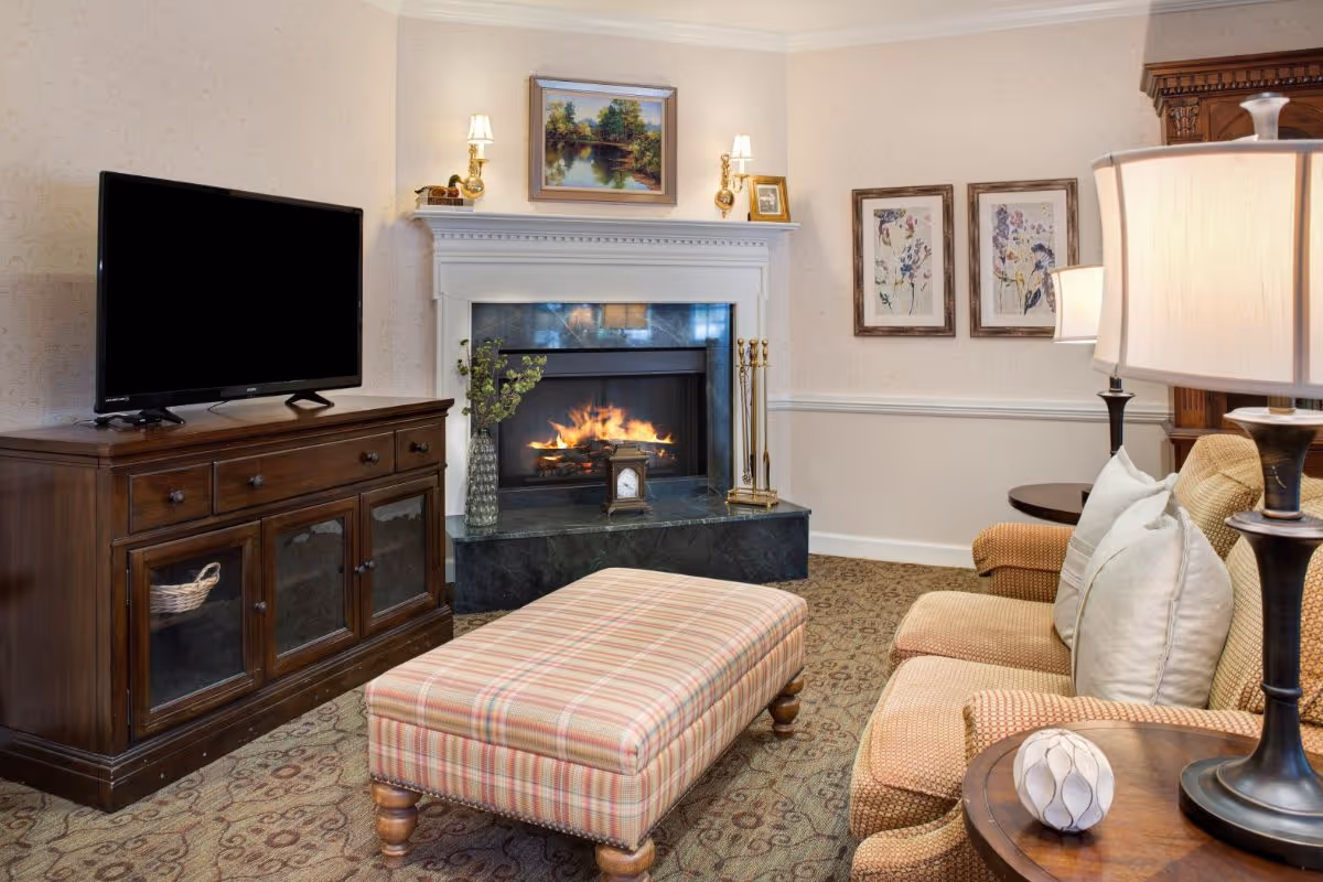 Cozy living room with a lit fireplace, a flat-screen TV on a wooden cabinet, a plaid upholstered ottoman, and a beige sofa with white pillows. The room is decorated with framed artwork, table lamps, and a patterned carpet.