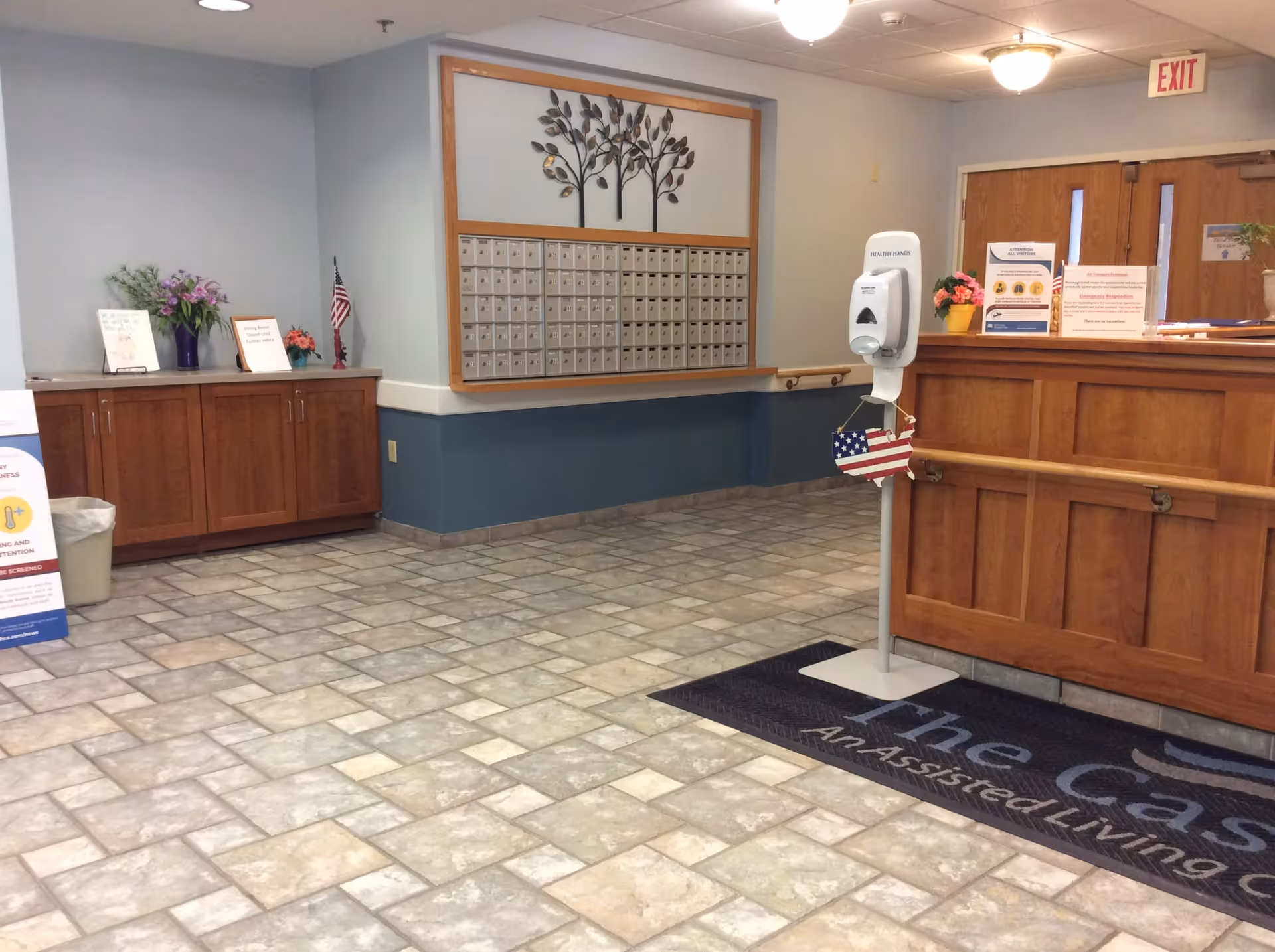 Interior view of a reception area in an assisted living facility with a wooden front desk, a hand sanitizer stand, a wall of mailboxes, and cabinets with flowers and signs on top. The floor is tiled, and there is an exit door in the background.