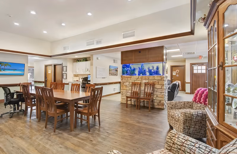 Bright senior living common room with a large wooden dining table and chairs, an aquarium set in a stone divider, and seating area with armchairs.