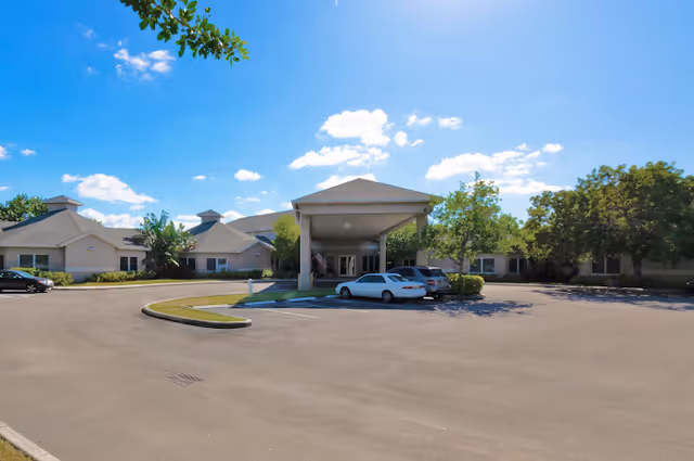 Exterior view of a single-story senior living facility with a covered entrance and a parking area with several cars. The building is surrounded by trees and landscaping under a blue sky with scattered clouds.