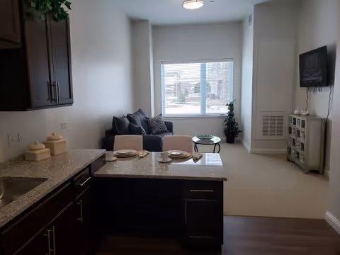 Open-plan apartment interior with a granite kitchen island in the foreground and a small living area with a sofa, TV, and window in the background.