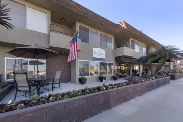 Exterior view of Brookdale Valley View senior living facility showing a two-story building with balconies, an American flag, outdoor seating with umbrellas, and landscaped flower beds along the sidewalk.