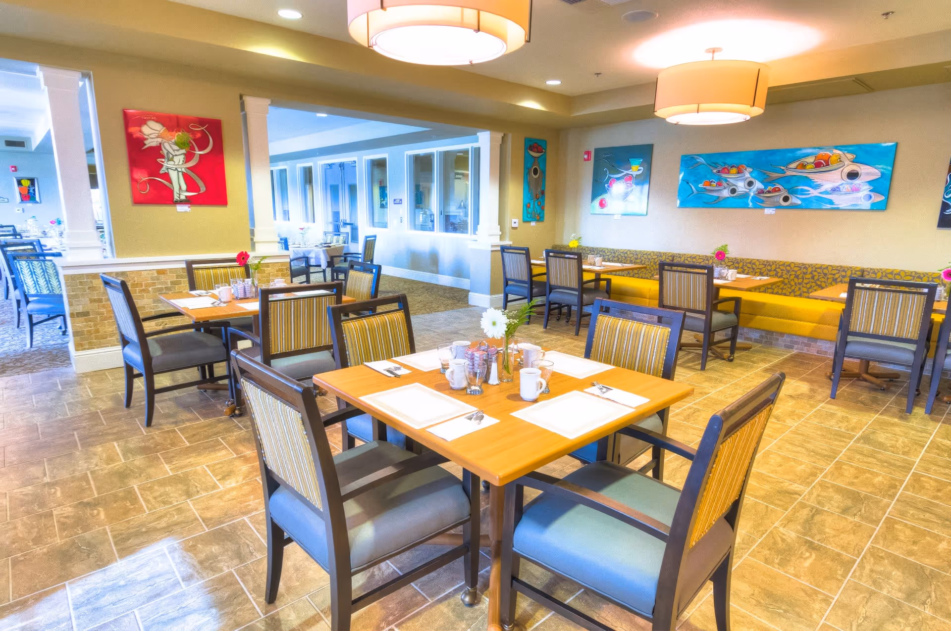 A bright and clean dining room with multiple wooden tables and cushioned chairs arranged neatly. Each table is set with placemats, cups, and utensils, and decorated with small flower vases. The walls are adorned with colorful abstract paintings, and there are large ceiling lights providing warm illumination. The floor is tiled, and there is a yellow cushioned bench along one wall.