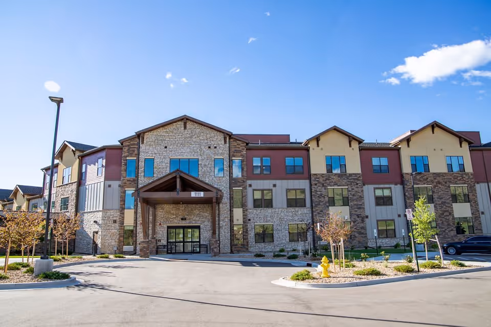 Three-story assisted living building with a stone and siding facade and covered main entrance under a clear blue sky.