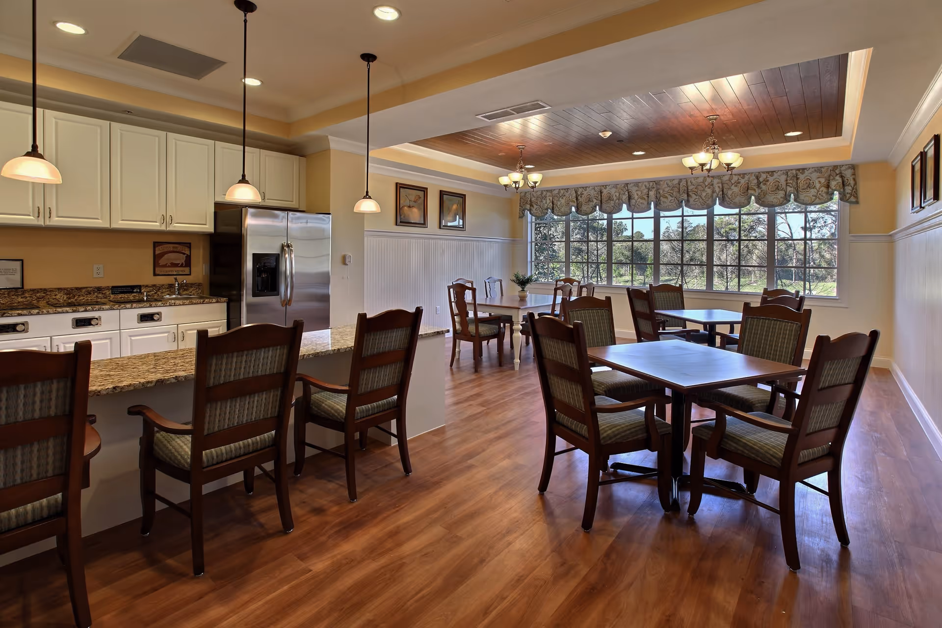 A bright dining area in a senior living facility featuring wooden floors, multiple tables with chairs, a kitchen area with white cabinets, granite countertops, stainless steel refrigerator, and pendant lighting. Large windows with decorative valances allow natural light to fill the room.