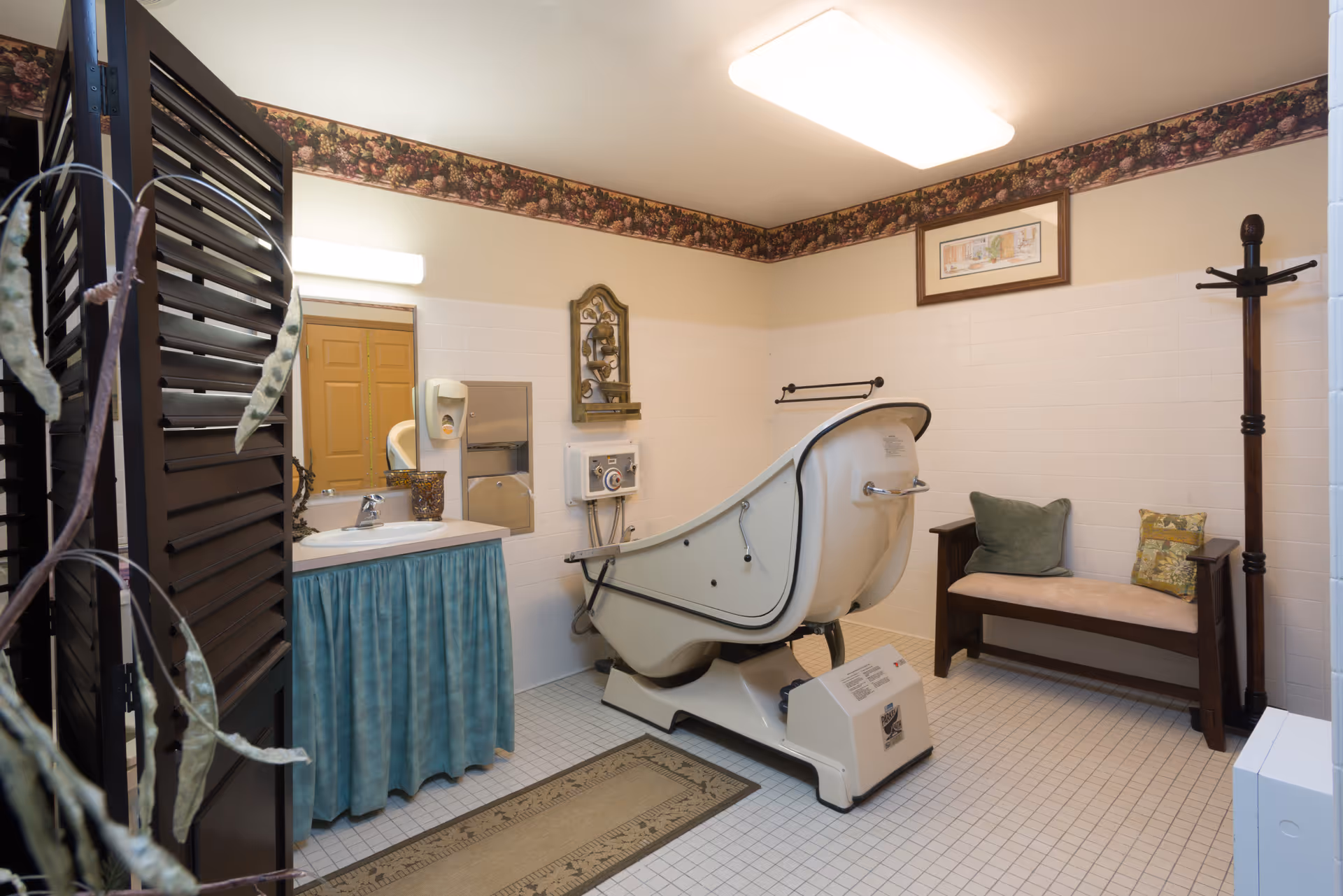 A vintage-style bathroom with a large white bathtub equipped with mechanical controls, a sink with a blue skirted cabinet, a wall-mounted soap dispenser, a wooden bench with two cushions, a coat rack, and floral wallpaper border near the ceiling.