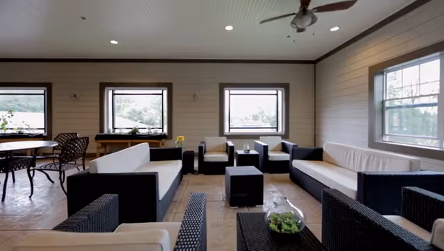 A bright and spacious indoor lounge area with multiple black wicker sofas and armchairs featuring white cushions. There are small black tables between the seating, one with a glass bowl containing green plants. The room has light-colored walls with horizontal paneling, three windows letting in natural light, a ceiling fan, and recessed lighting.