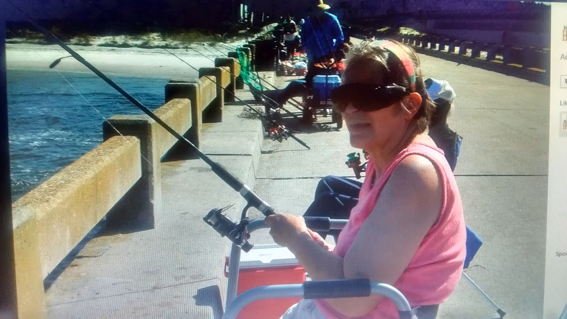 An elderly woman wearing sunglasses and a pink sleeveless top is sitting on a chair by a waterfront, holding a fishing rod. Other people are also fishing along the concrete pier with fishing rods lined up against the railing. The water and sandy shore are visible in the background.