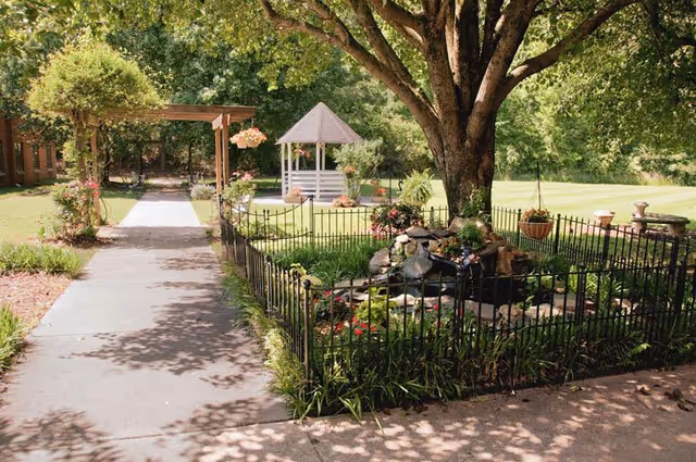 A peaceful outdoor garden area with a large tree providing shade over a small fenced garden featuring a water fountain. A paved walkway leads to a wooden pergola and a white gazebo in the background, surrounded by green grass and various plants and flowers.