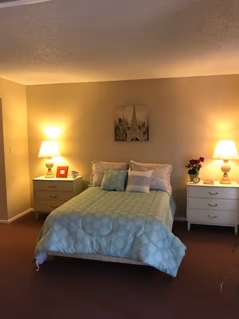 A cozy bedroom with a bed centered against a beige wall. The bed is made with a light blue patterned comforter and several pillows. On each side of the bed are white nightstands with drawers, each holding a lit table lamp. The left nightstand also has a small framed picture and a cup, while the right nightstand has a vase with red flowers and a small decorative item. Above the bed hangs a framed picture featuring a cityscape with the Eiffel Tower. The room has a warm, inviting ambiance.