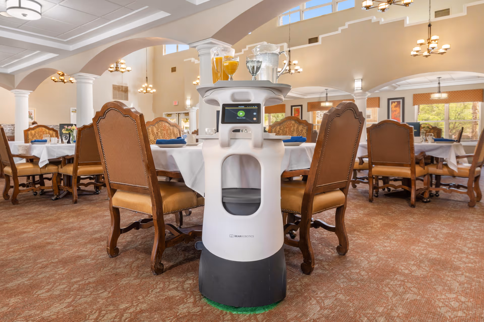 A dining room in an assisted living facility with multiple tables and chairs arranged neatly. In the center foreground, there is a white service robot carrying a tray with glasses of orange juice and water. The room has high ceilings, arched columns, and large windows allowing natural light to enter.