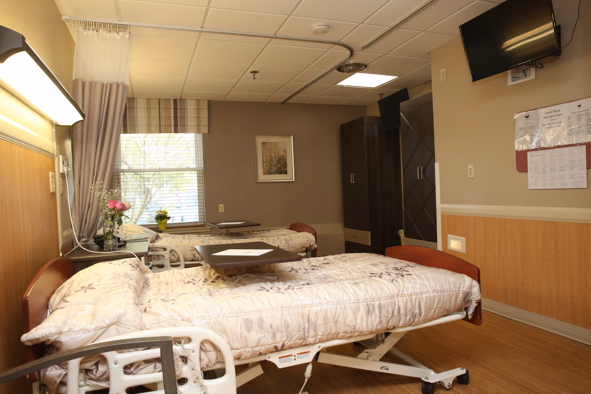 A room with two hospital-style beds with floral patterned bedding, each equipped with an overbed table. There is a window with blinds and curtains, a small table with flowers, a wall-mounted TV, and a bulletin board with papers on the wall. The room has wood paneling on the lower half of the walls and a light brown color scheme.