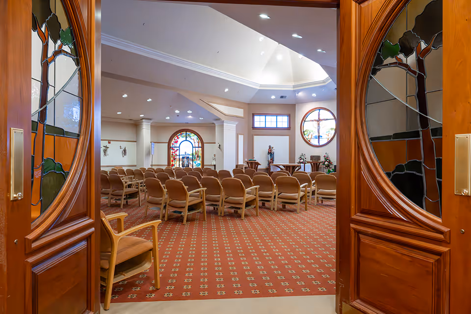 View through ornate wooden double doors into a chapel-style meeting room with rows of chairs and stained-glass windows.