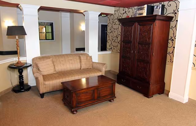 Small furnished lounge with a striped sofa, wooden coffee table, tall carved armoire, side table lamp, and decorative columns.