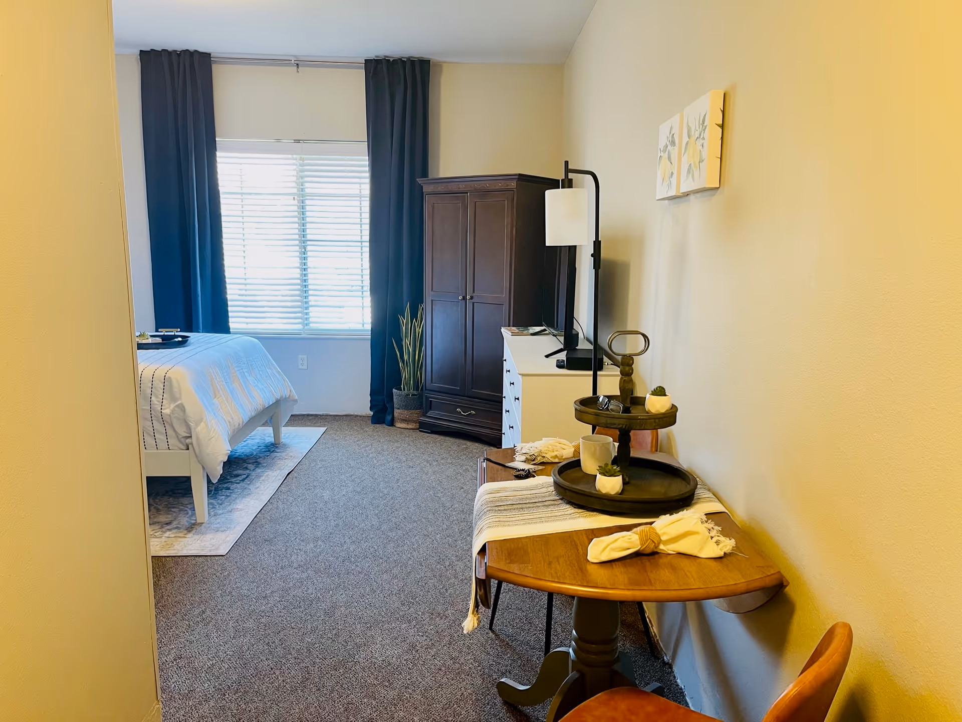 A cozy bedroom in AVIVA Merrillville featuring a bed with white bedding near a window with dark curtains. There is a dark wooden wardrobe and a white dresser with a lamp on top. A small wooden table with a two-tiered tray, a cup, and decorative items is placed against the wall, accompanied by a chair. The walls are painted light beige and the floor is carpeted.